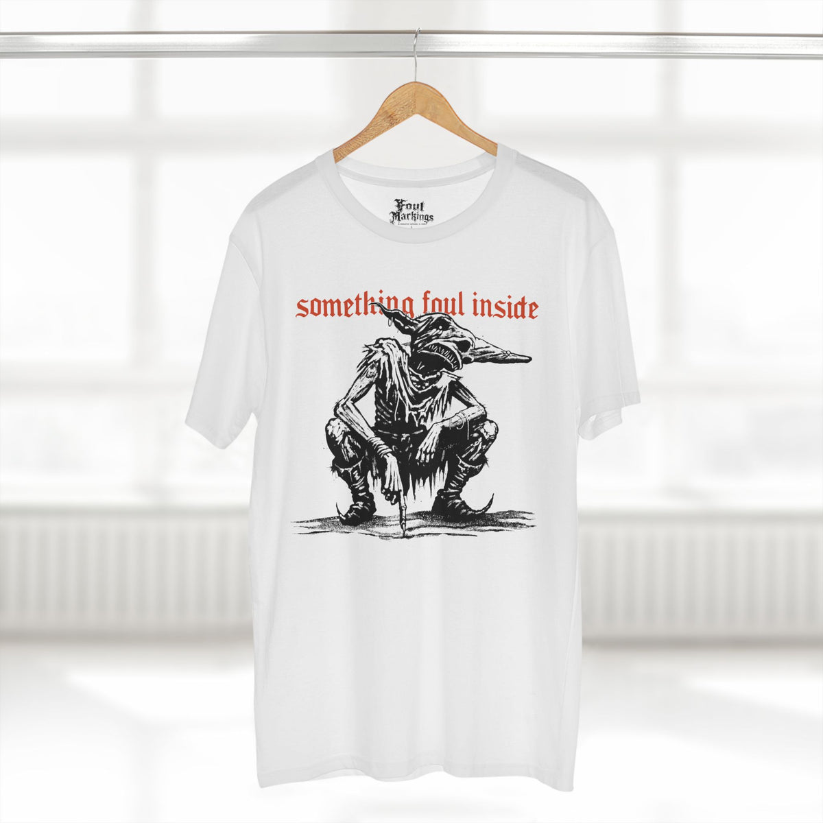 White tee with a black ink crouching goblin under red gothic text reading something foul inside, shown hanging
