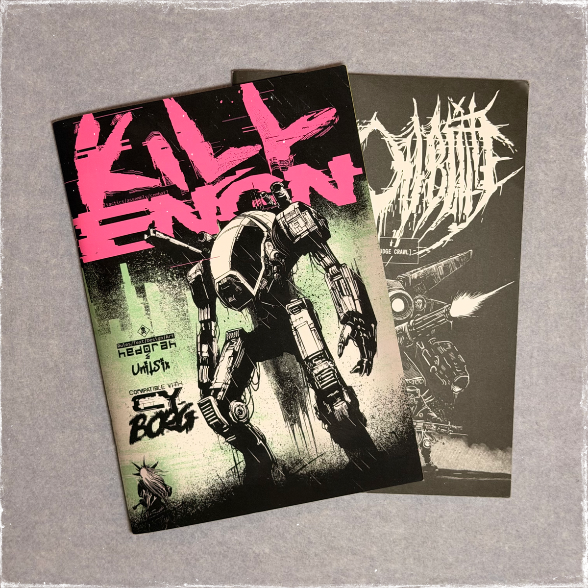 Both zine covers fanned out, a pink and green mech on Kill ENGN over the Cast Oubliette cover